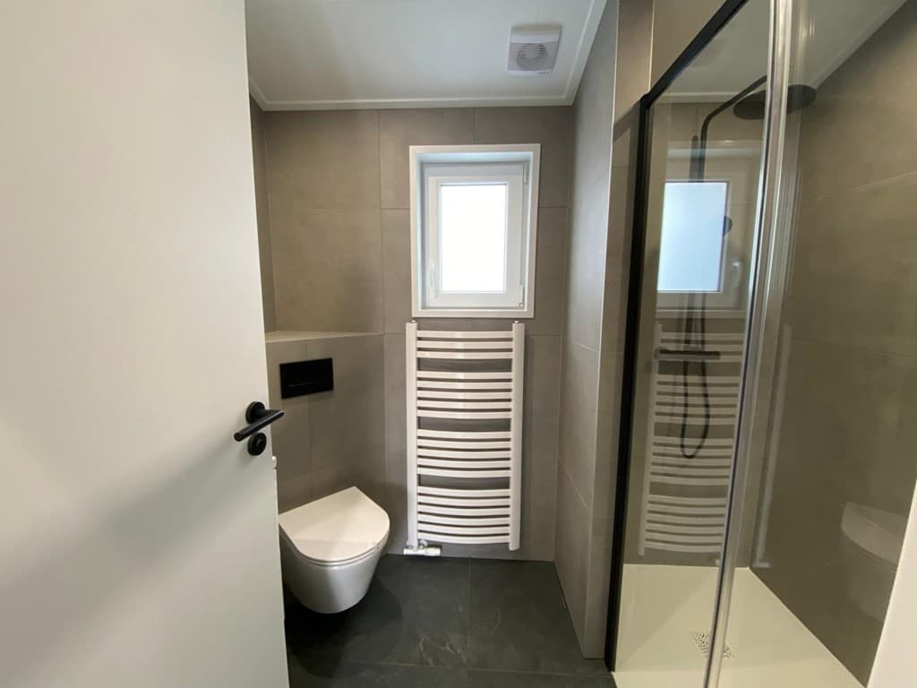 Moderne badkamer met douche, toilet en handdoekenradiator, afgewerkt met tegelwerk en een raam voor natuurlijke lichtinval
