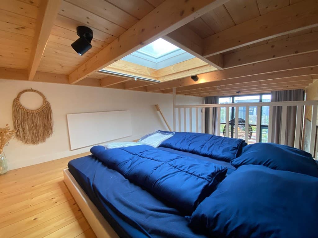 Sfeervolle slaapkamer met houten plafond, groot dakraam en tweepersoonsbed met blauwe dekbedden.