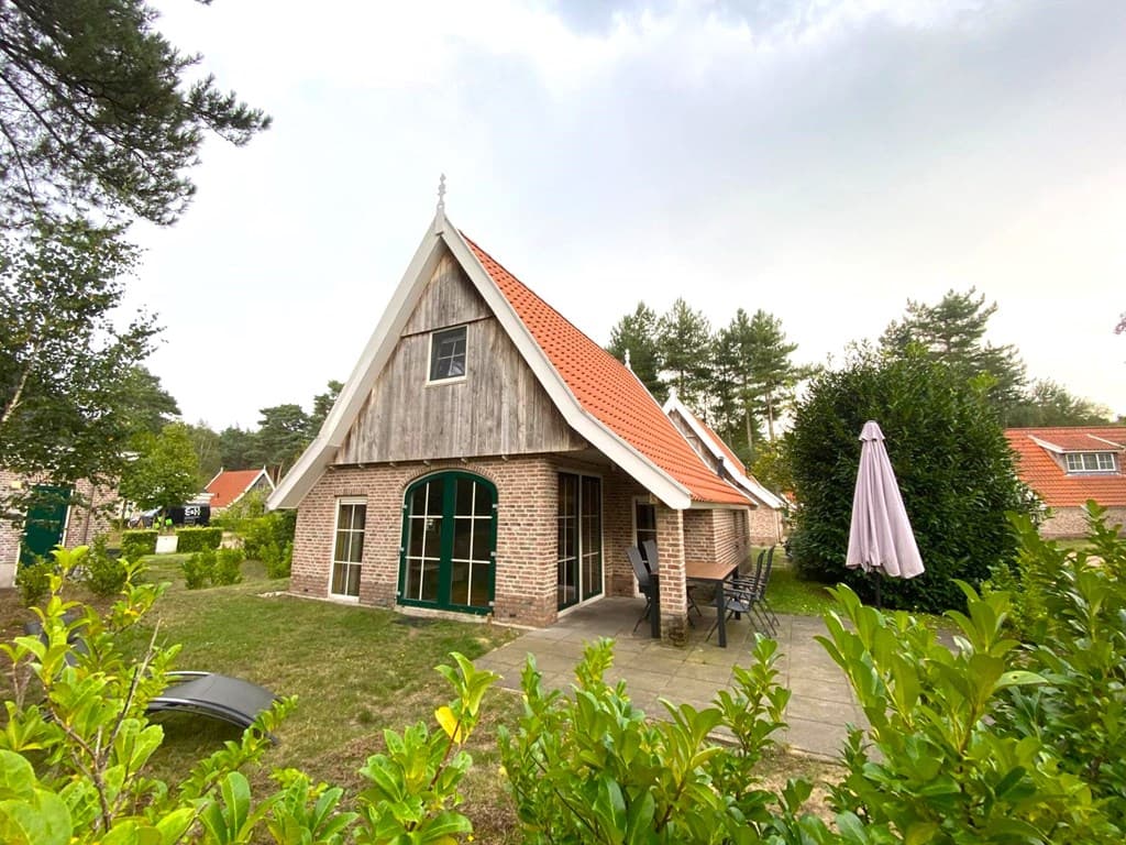 Vakantiehuis met houten gevel en rode dakpannen, omgeven door groen, met terras en parasol aan de achterkant