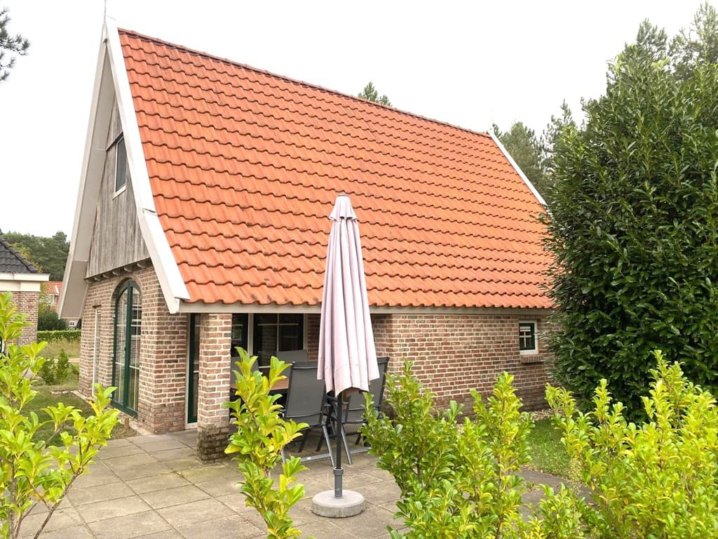 Vakantiewoning met rood pannendak, zicht op terras met parasol en omliggende groene beplanting.