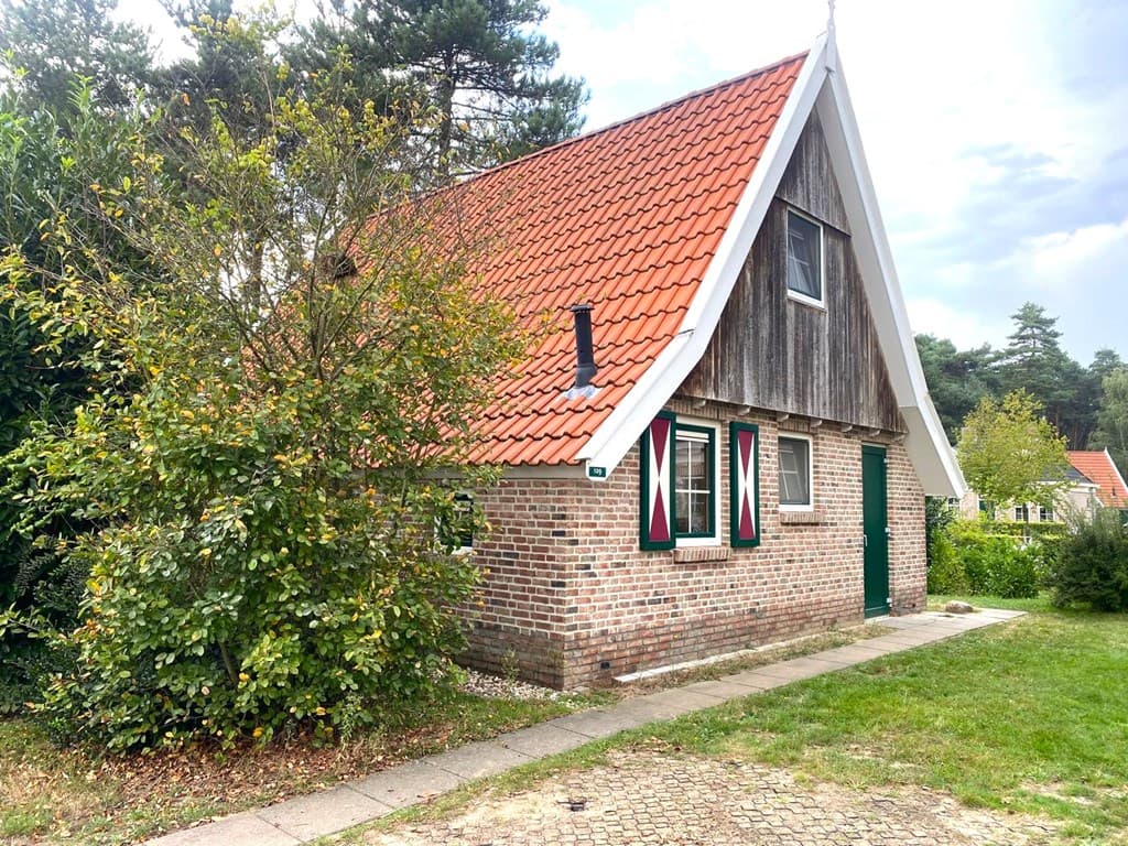 Vakantiehuis met rode dakpannen, houten gevel en groene deur, omringd door gras en struiken op Landal Landgoed De Hellendoorn