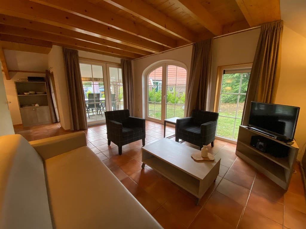 Gezellige woonkamer met houten balken, twee fauteuils, een bank, een tv en grote ramen naar de tuin.