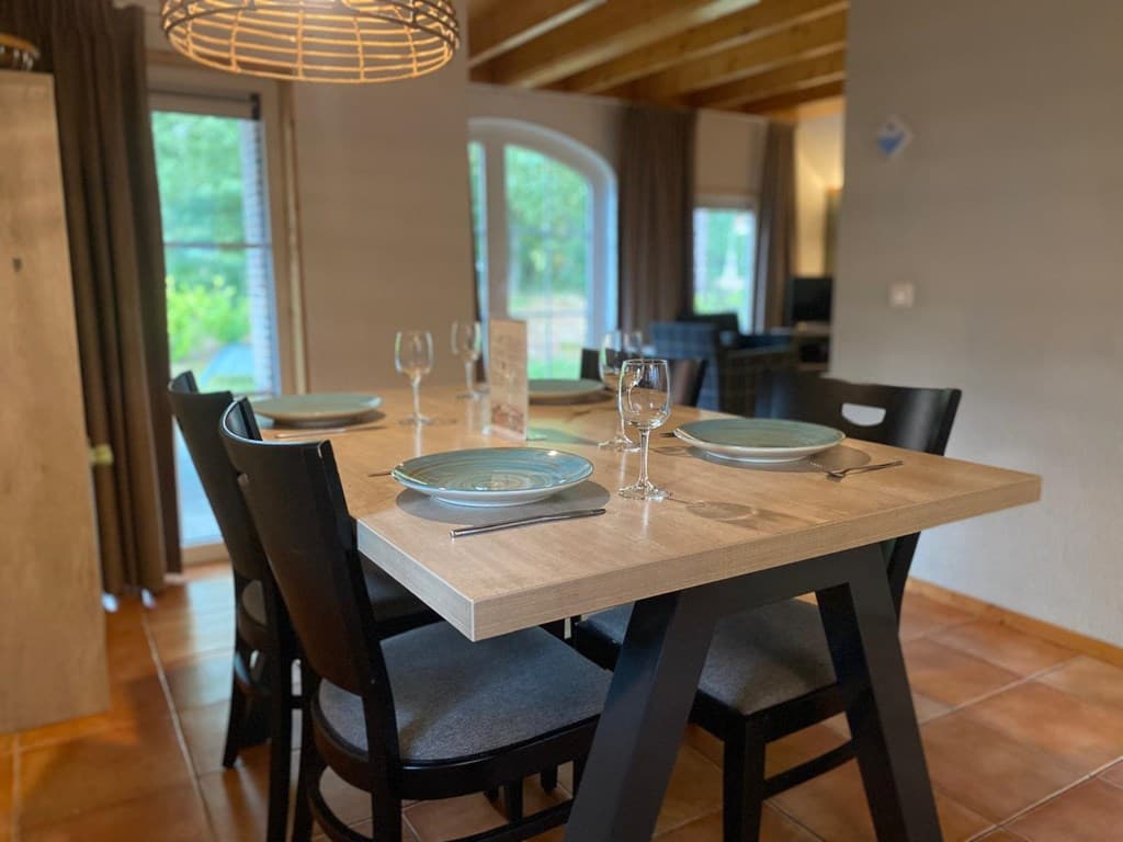 Eettafel met glazen en borden, vier stoelen, en een zicht op een groene tuin door de ramen.