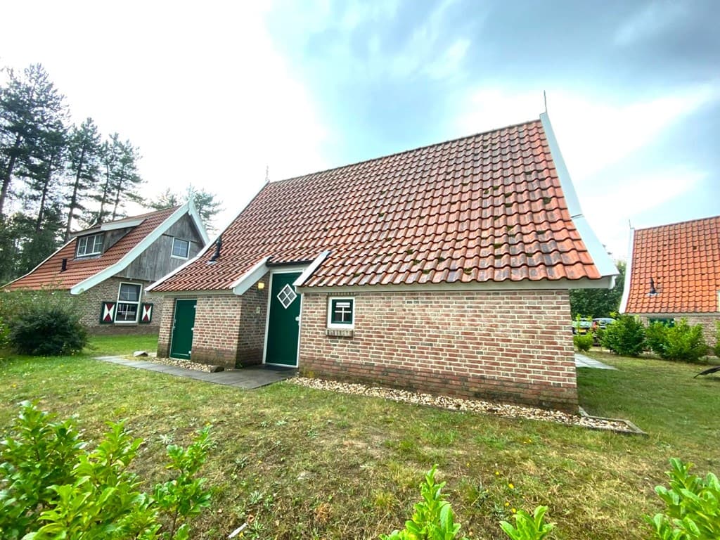 Klein vakantiewoning met rood-dak pannen, verhard pad en groen grasveld omringd door bomen en andere woningen.