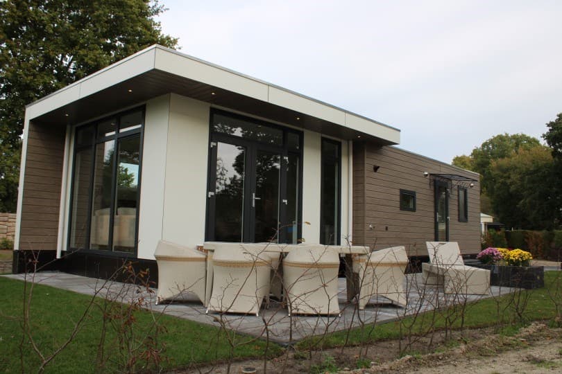 Moderne vakantiewoning met een terras en witte tuinstoelen, omgeven door groen en aangelegde plantenbedden.