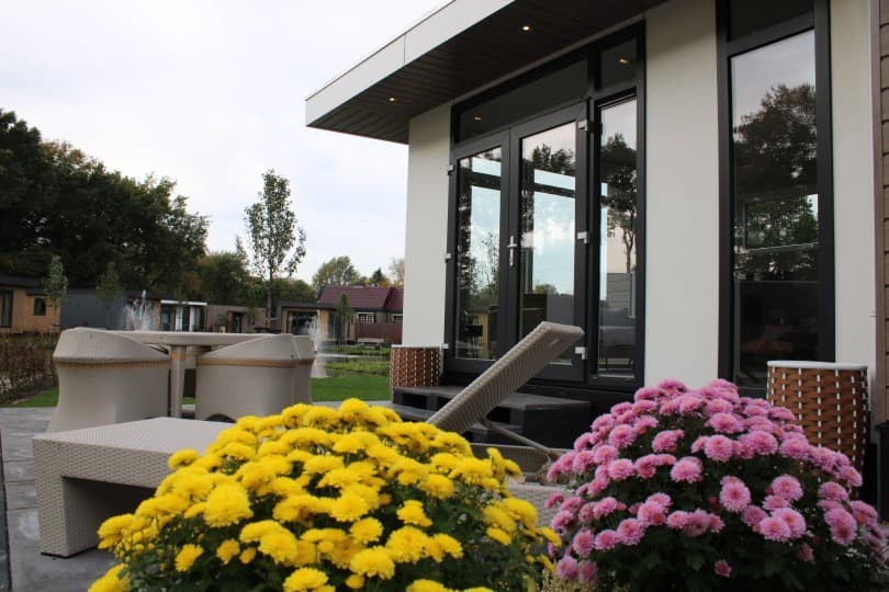 Moderne vakantiewoning met grote ramen, terrasmeubels en kleurrijke bloemen in de tuin.