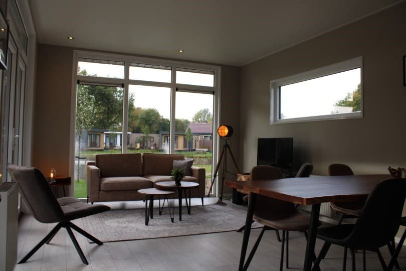 Ruime woonkamer met een bank, eettafel, lamp, tv en grote ramen met uitzicht op de tuin en andere woningen.