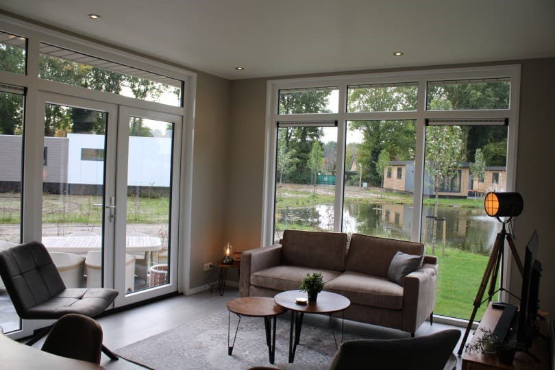 Moderne woonkamer met grote ramen, bank, salontafels en uitzicht op het water en de natuur buiten.