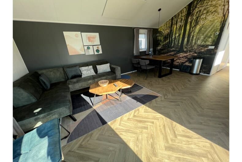 Woonkamer met een groene bank, houten tafel en grote natuurwanddecoratie, voorzien van een lichtinval vanuit een raam.