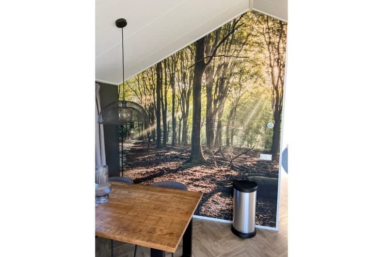 Binnenruimte met een houten tafel en een muur met een groot bosdecor, verlichting valt door de bomen