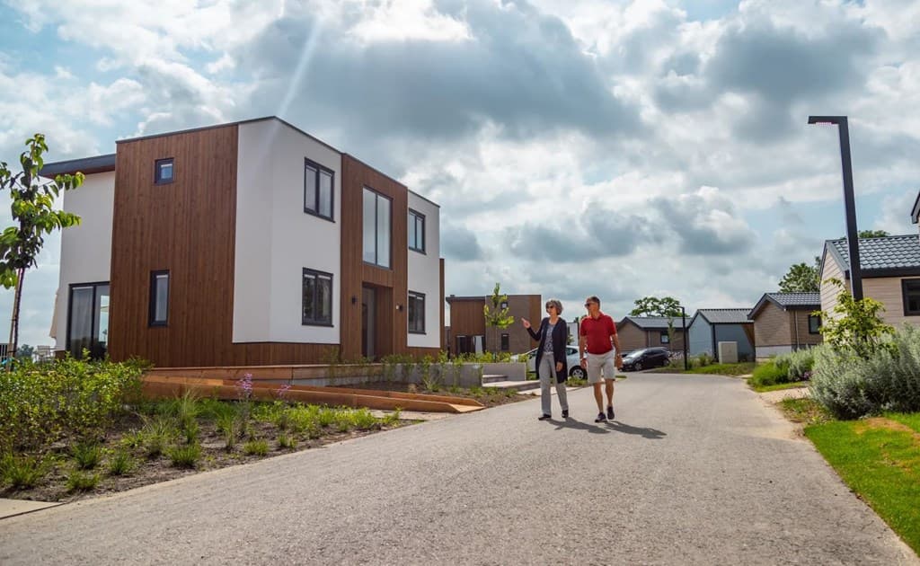 Twee mensen wandelen op een rustige weg langs moderne vakantiewoningen met groene beplanting en een heldere lucht.