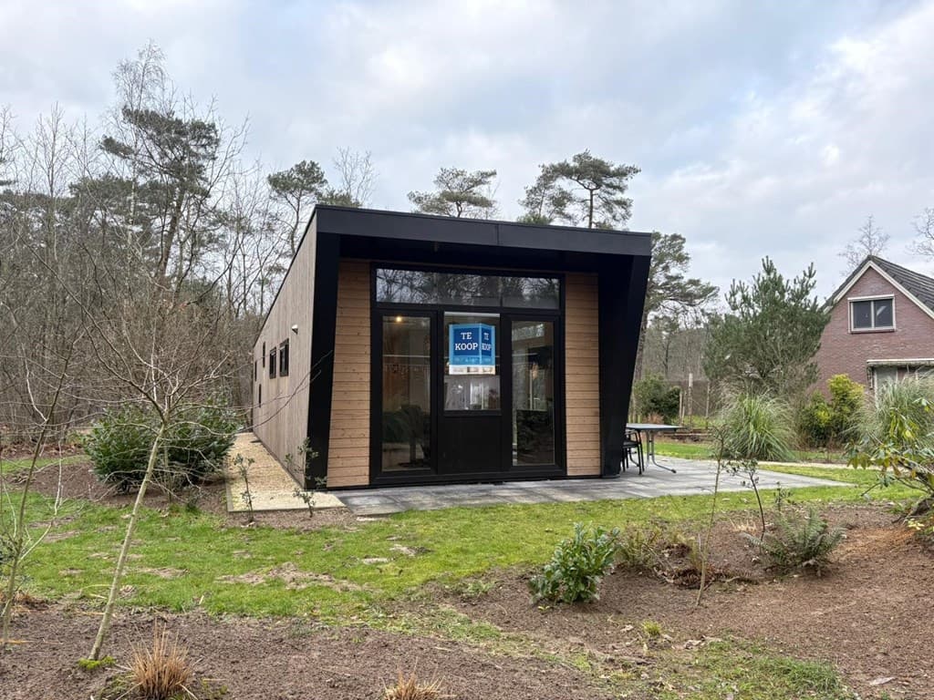 Moderne woning met zwarte gevel en grote glazen deuren, gelegen in een groene omgeving met bomen en gras.