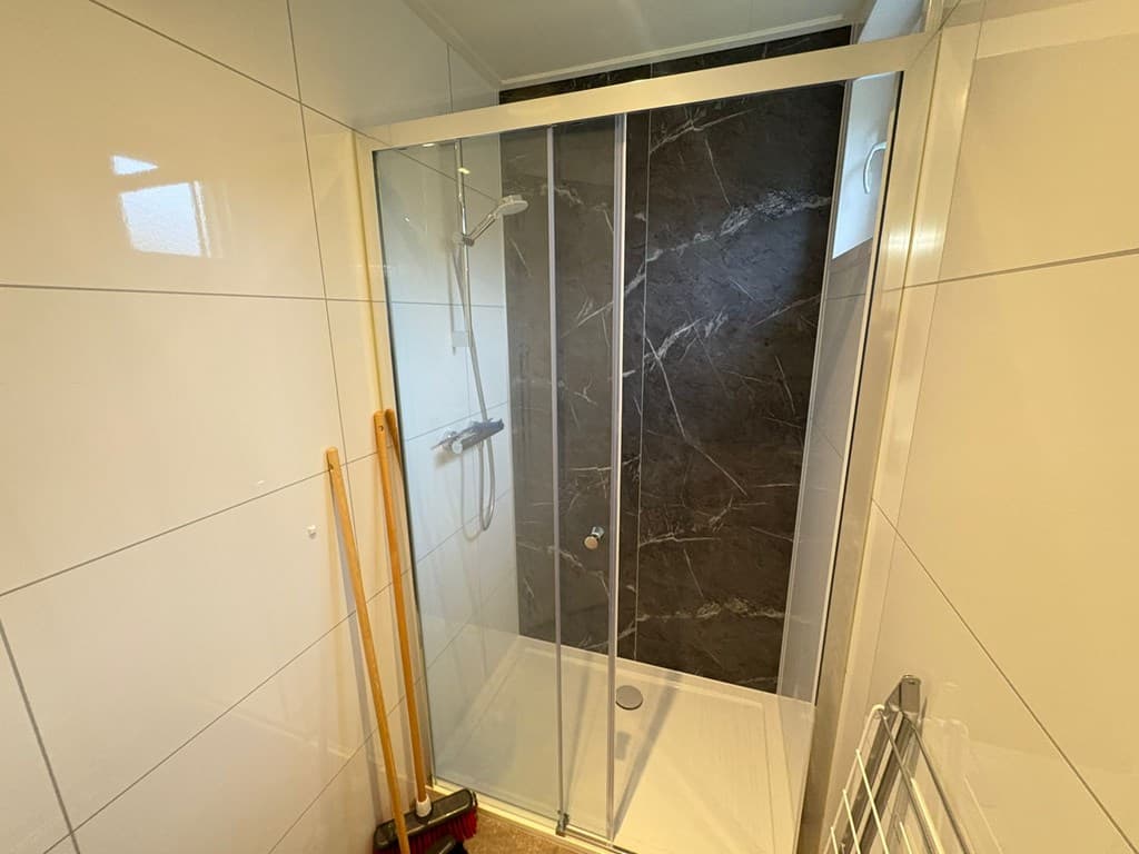Moderne douchecabine met glazen deuren en donkere wandplaten, naast schoonmaakmaterialen in de hoek