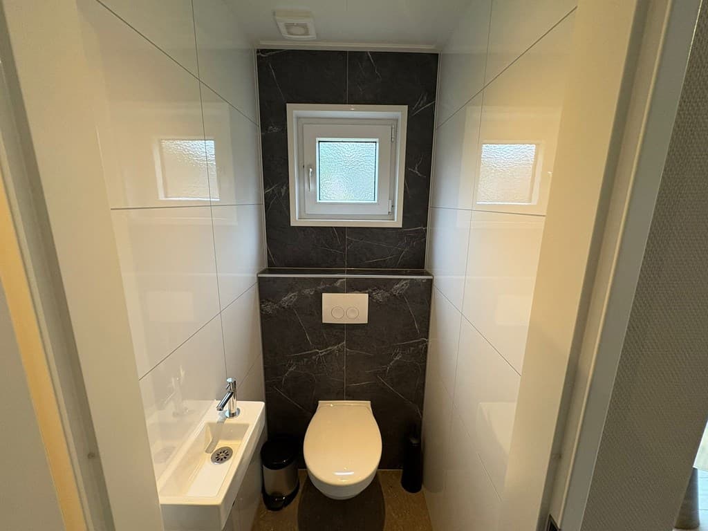 Modern toilet met witte wandtegels en een donkere achterwand, voorzien van een raam en een kleine wastafel.