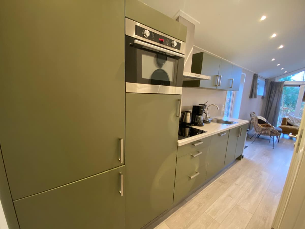 Keuken met groene kasten, oven, koffiezetapparaat en uitzicht op buiten via grote ramen.
