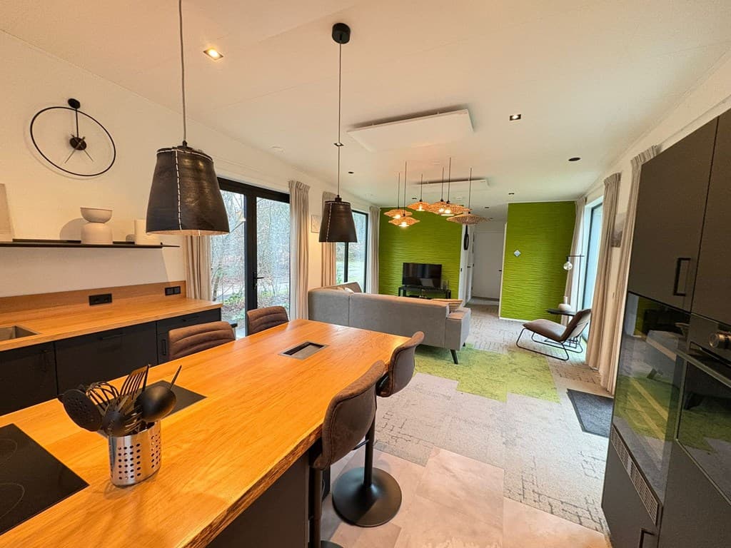 Moderne open keuken met houten tafel en groene accentwand, gezellige zithoek met televisie en grote ramen.