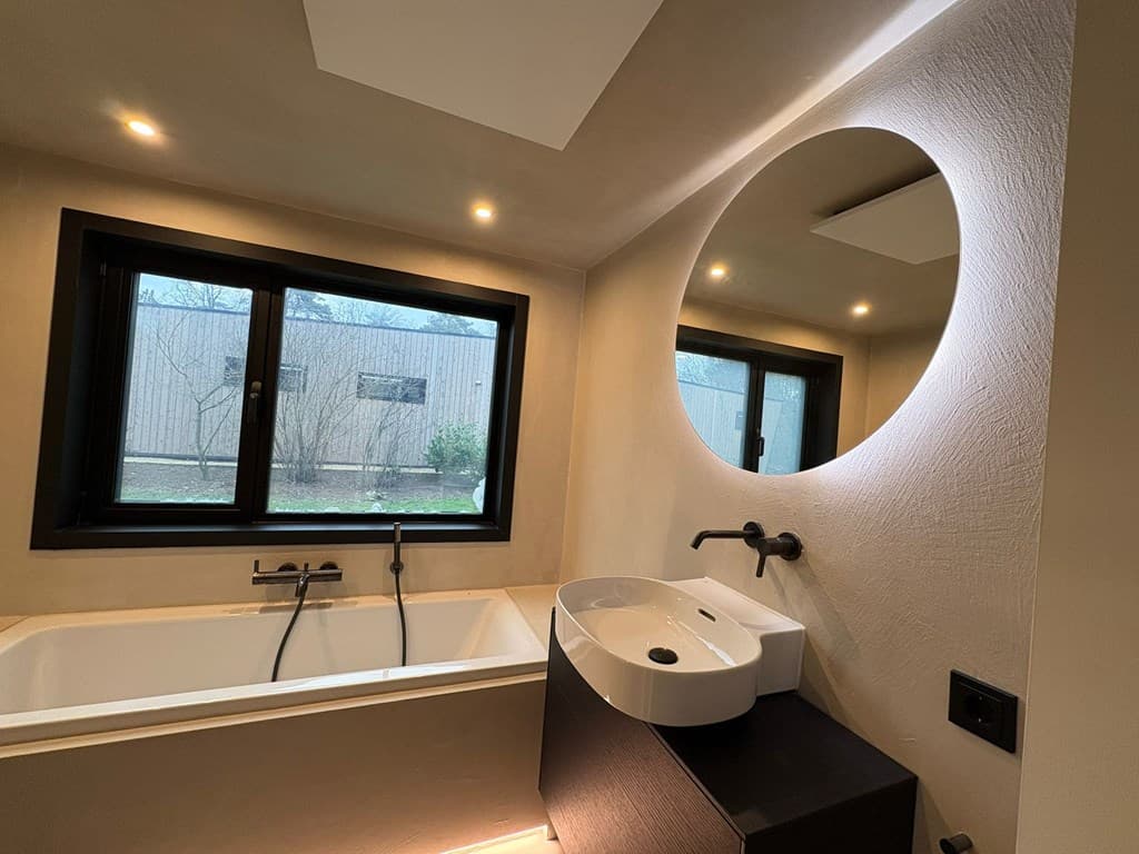 Moderne badkamer met bad, wastafel en grote ronde spiegel, twee ramen met uitzicht op de tuin.
