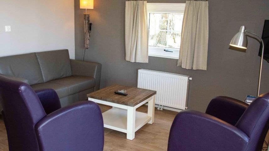Zithoek met grijze bank, paarse stoelen en houten salontafel bij een raam met gordijnen.