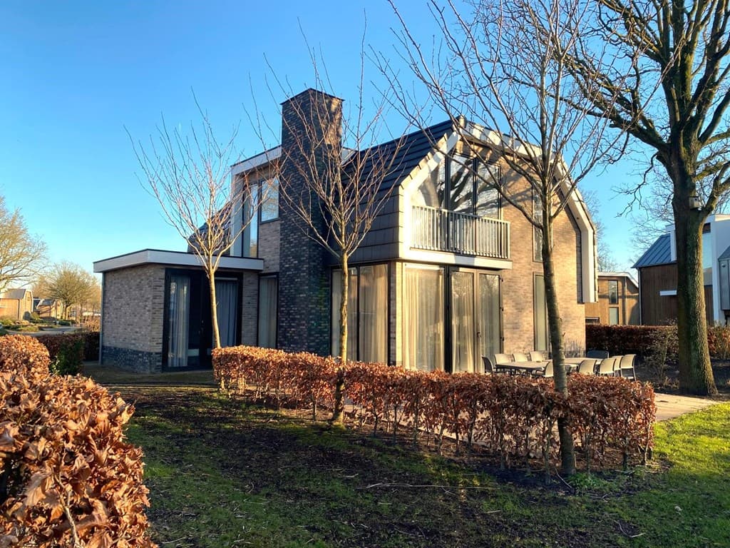 Twee onder één kap woning met moderne gevel, grote ramen en een terras, omringd door een laag haagje en bomen.