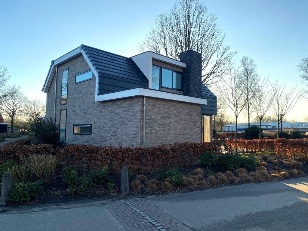 Moderne vakantiewoning met een lichte stenen gevel, schuin dak en grote ramen, omgeven door een verzorgde tuin en bomen.