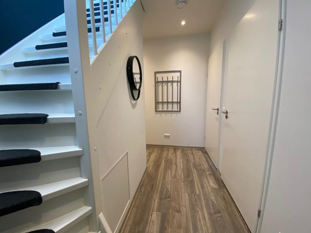 Ingang van de woning met een trap, muur met een spiegel en een schoenenrek, en een houten vloer.
