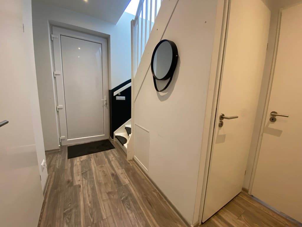 Lichte entree met een houten vloer, deur naar buiten, trap naar de bovenverdieping en een ronde spiegel aan de muur.