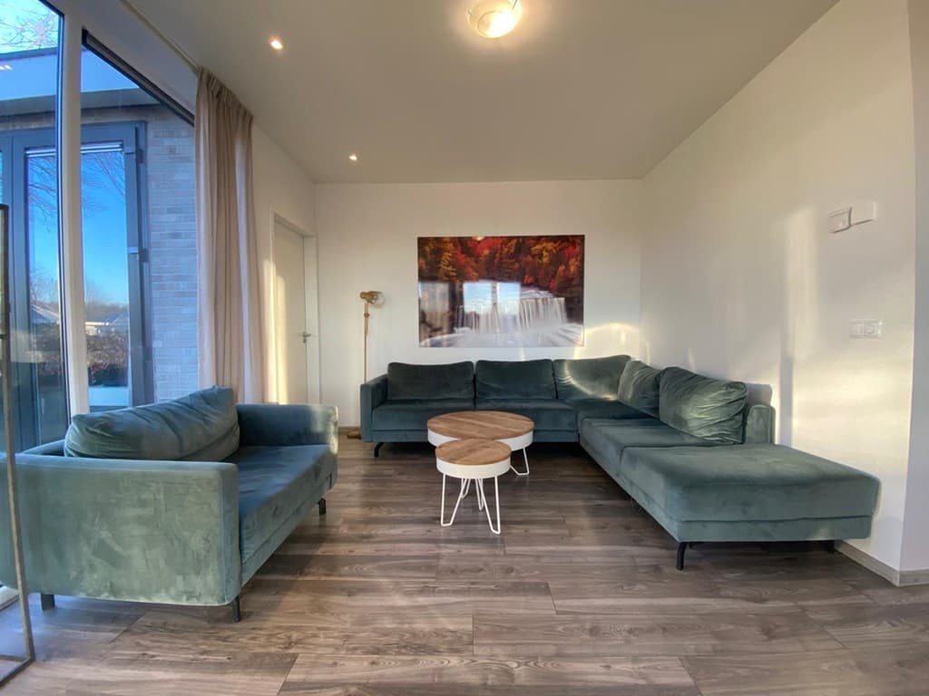 Lichte woonkamer met groene meubelen, een houten salontafel en een groot raam met uitzicht op buiten.