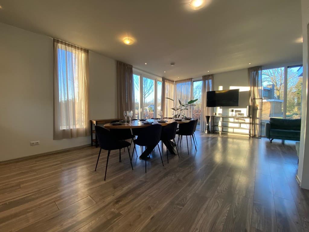 Ruime woonkamer met eettafel, moderne meubels en grote ramen met veel natuurlijk licht.