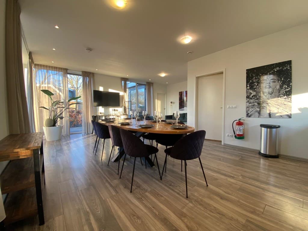 Ruimtelijke eetkamer met grote tafel, gezellige stoelen, open deur naar buiten en moderne inrichting.