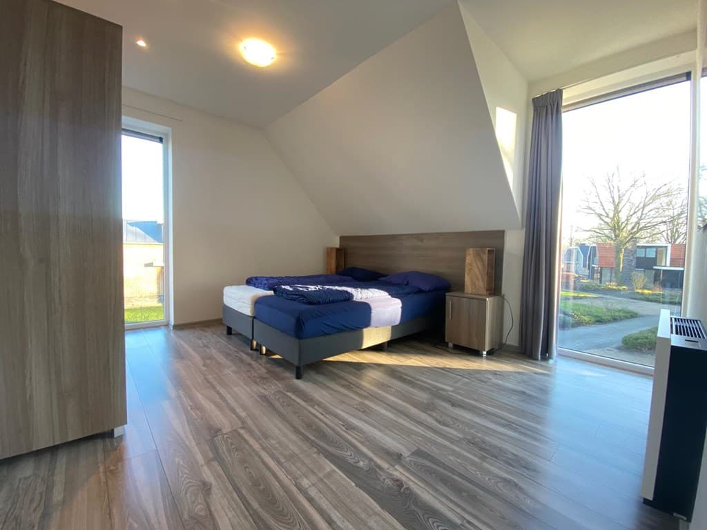 Slaapkamer met een tweepersoonsbed, houten vloer en grote raam met zicht op buiten.
