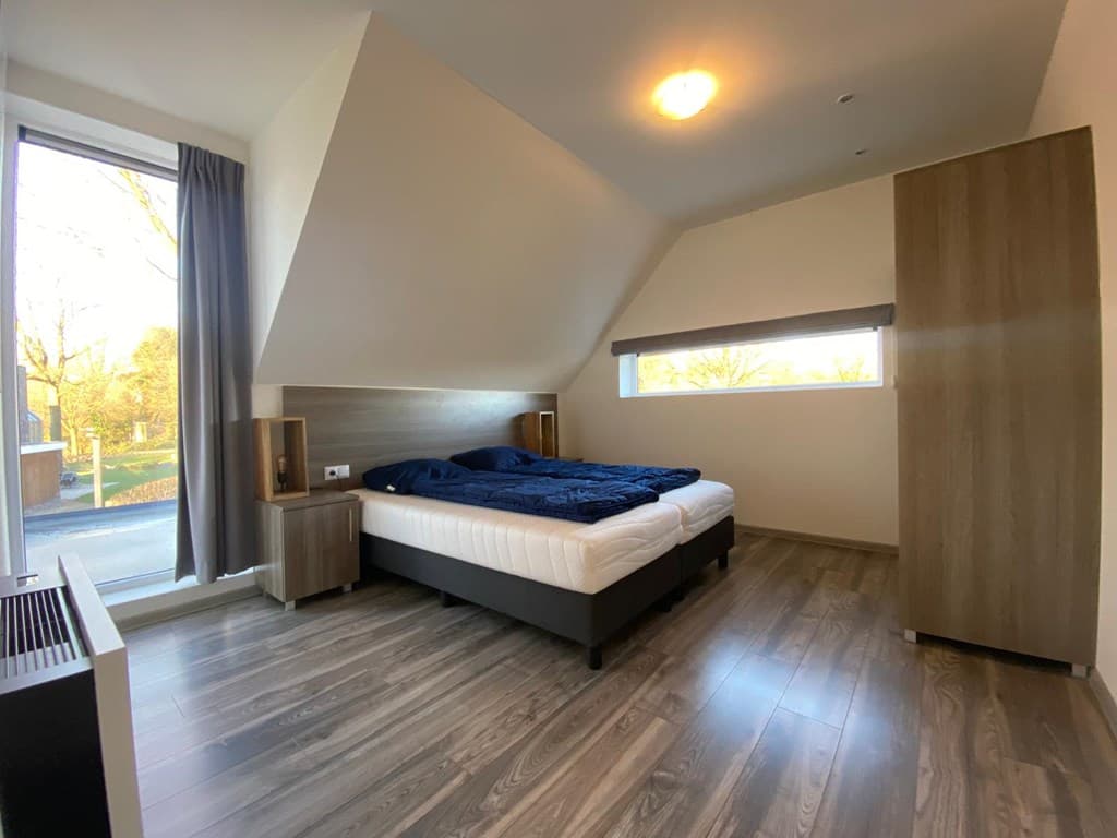 Slaapkamer met een groot raam, houten vloer, twee nachtkastjes en een kledingkast, moderne inrichting met blauwe dekbedden.