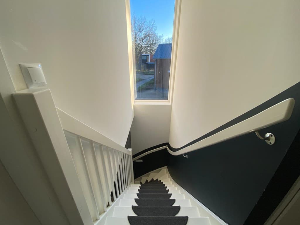 Trap met witte leuning en zwarte muur, uitzicht op een raam met natuurlijk licht dat de ruimte verlicht