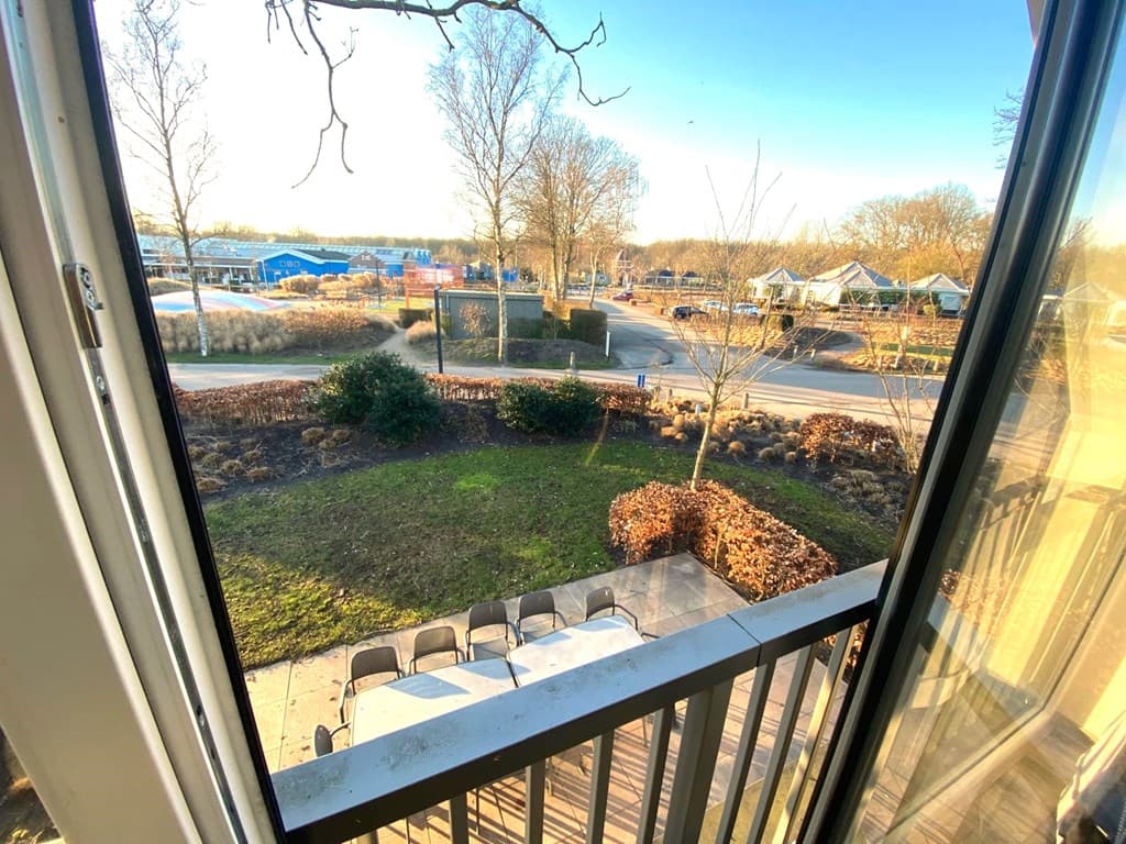 Uitzicht vanuit een woning met een tuin, grasveld en tafels, omringd door bomen en verkeersweg in de verte.