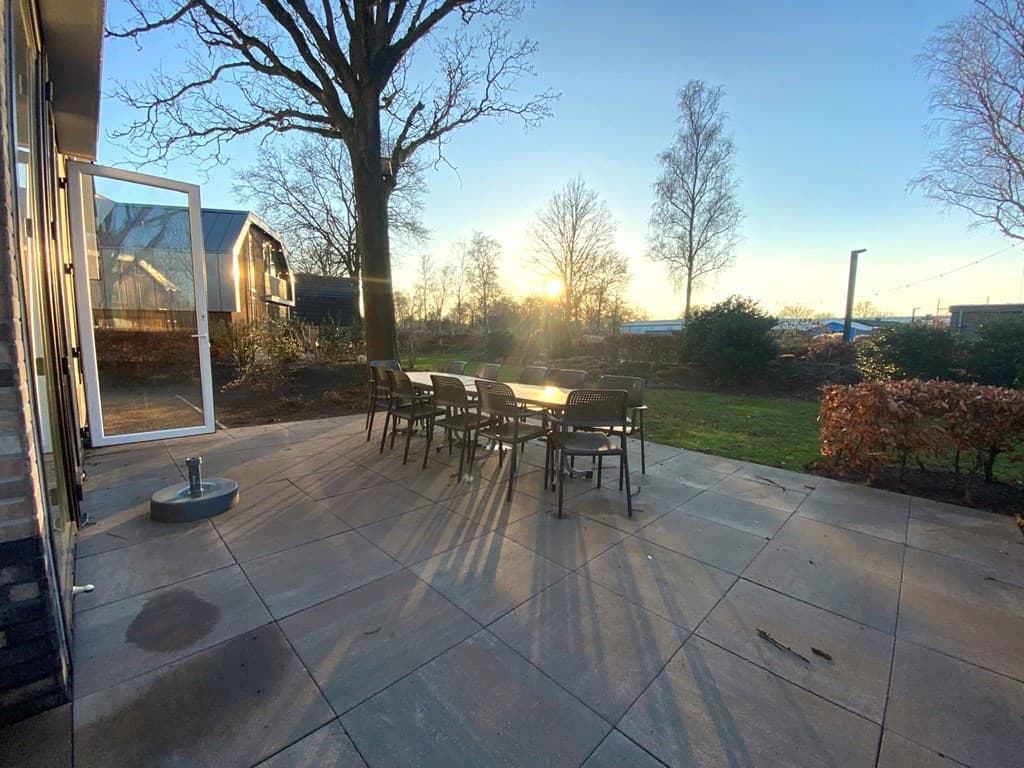 Terras met een tafel en stoelen, omringd door bomen en een grasveld, met de zon die ondergaat in de achtergrond