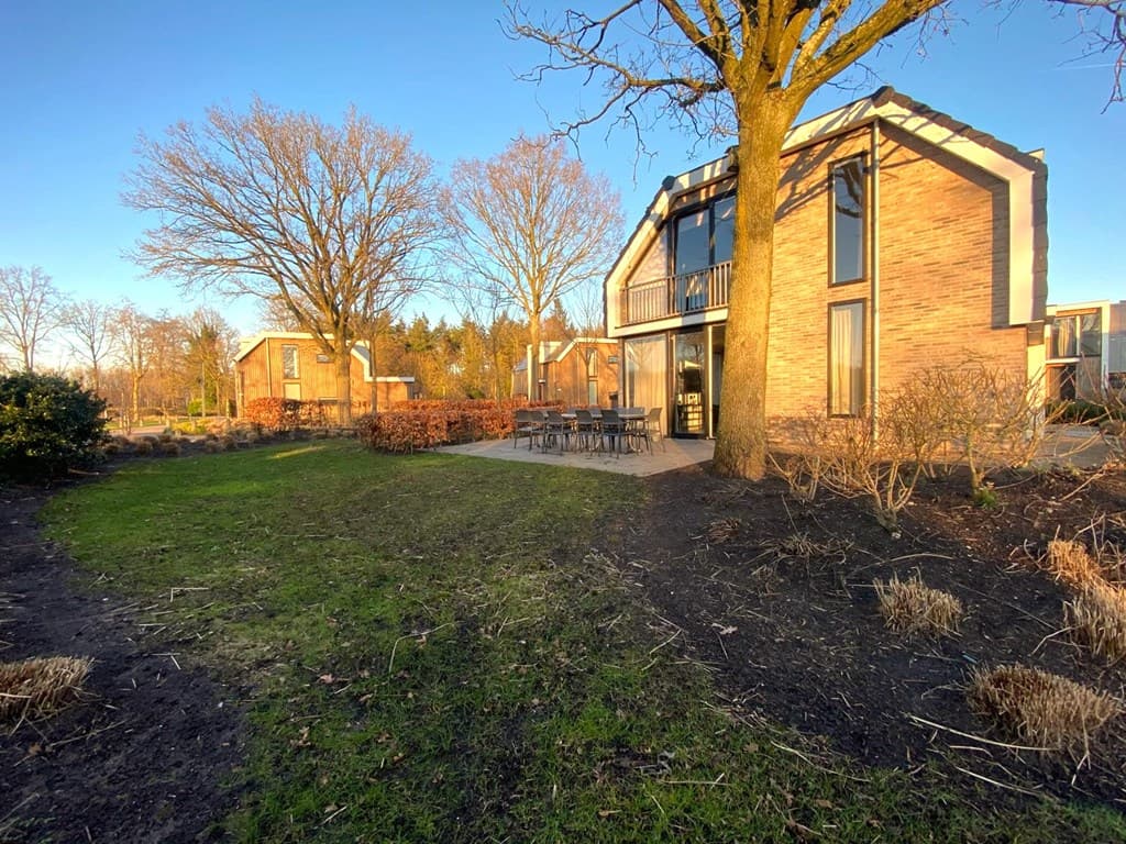 Twee moderne woningen met grote ramen en een terras in een groene tuin omgeven door bomen en struiken.