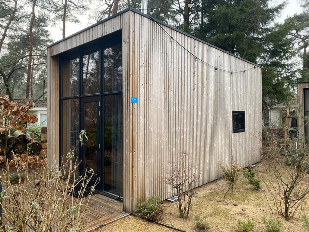Moderne houten woning met grote glazen gevel en omgeving van bomen en struiken.