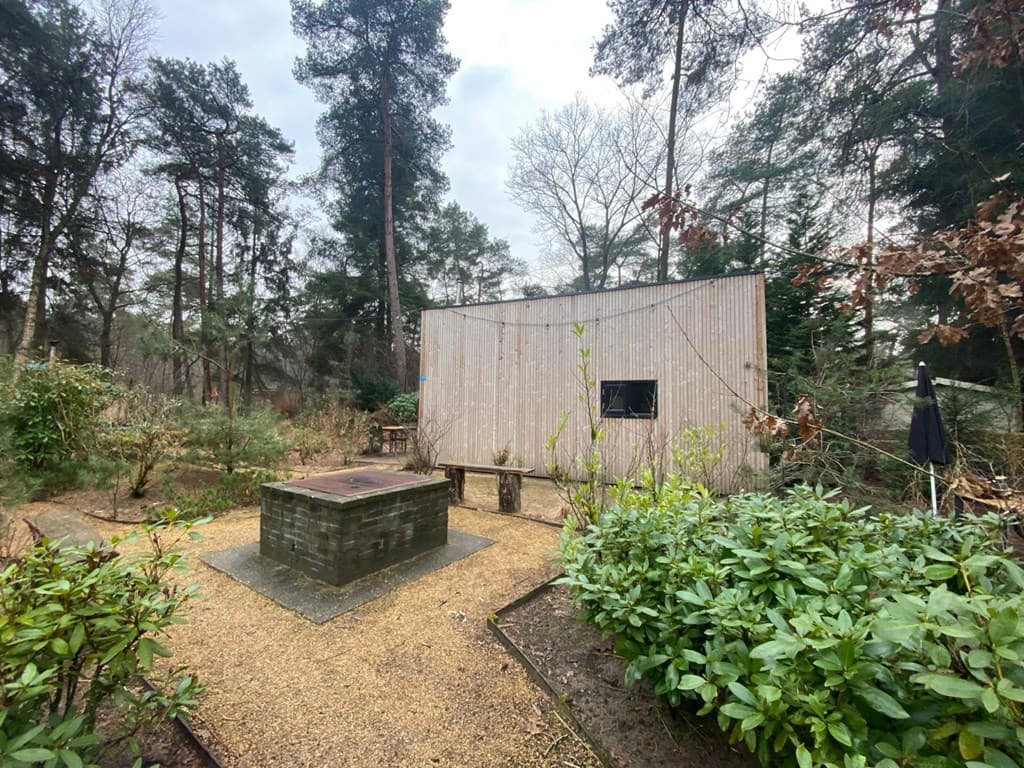 Houten vakantiewoning omringd door bomen en struiken, met een stenen barbecueplaats voor het huis.