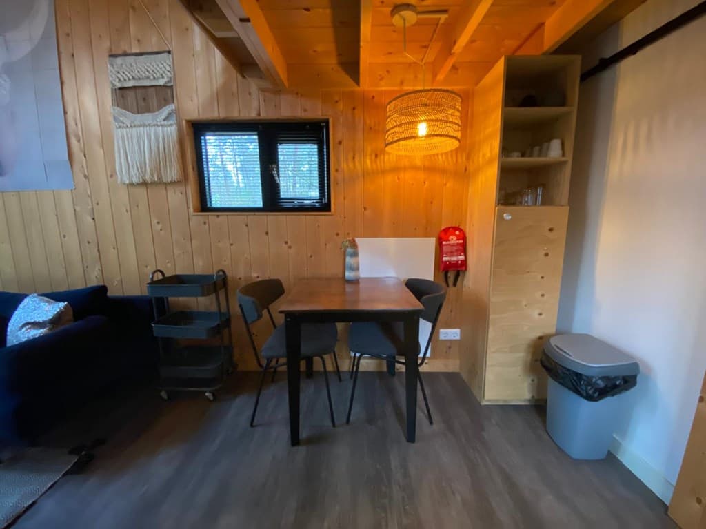 Houtachtige inrichting met een eettafel en stoelen, een bank, wandplank en een prullenbak zichtbaar in de ruimte