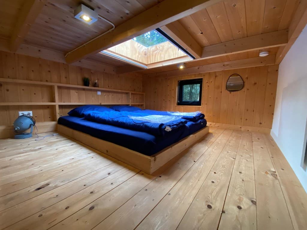 Slaapkamer met houten plafond, bed met blauw dekbed, kleine vensters en een lichtkoepel voor natuurlijk licht
