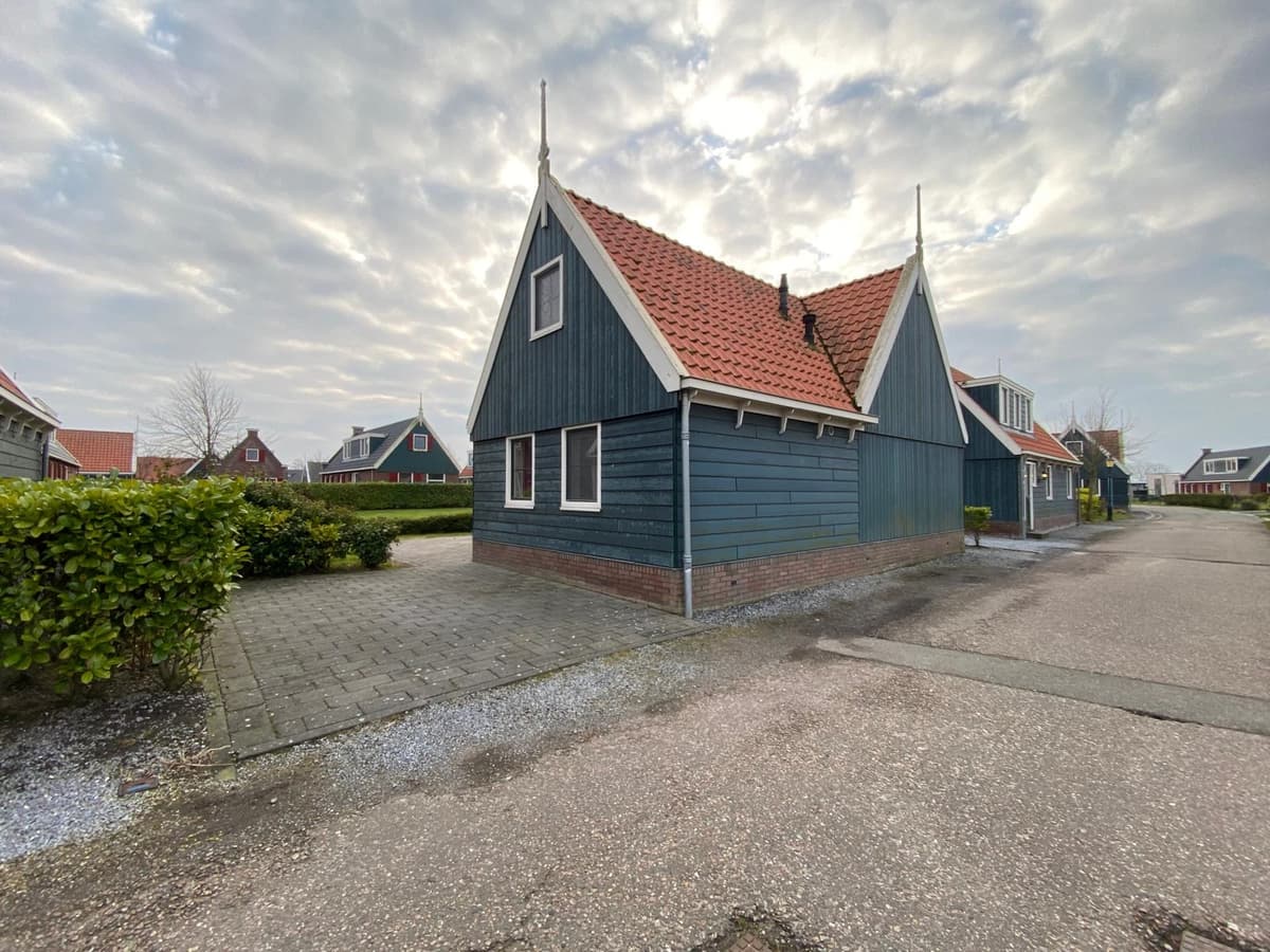 Houten vakantiehuis met een schilddak en rode pannen, gelegen aan een verhard pad met groenheken.
