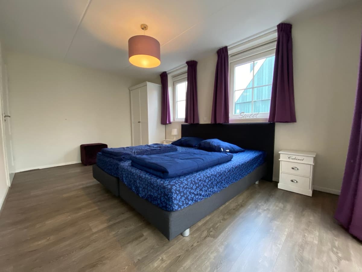 Slaapkamer met tweepersoonsbed, nachtkastje en kast; paarse gordijnen bij de ramen en laminaatvloer.