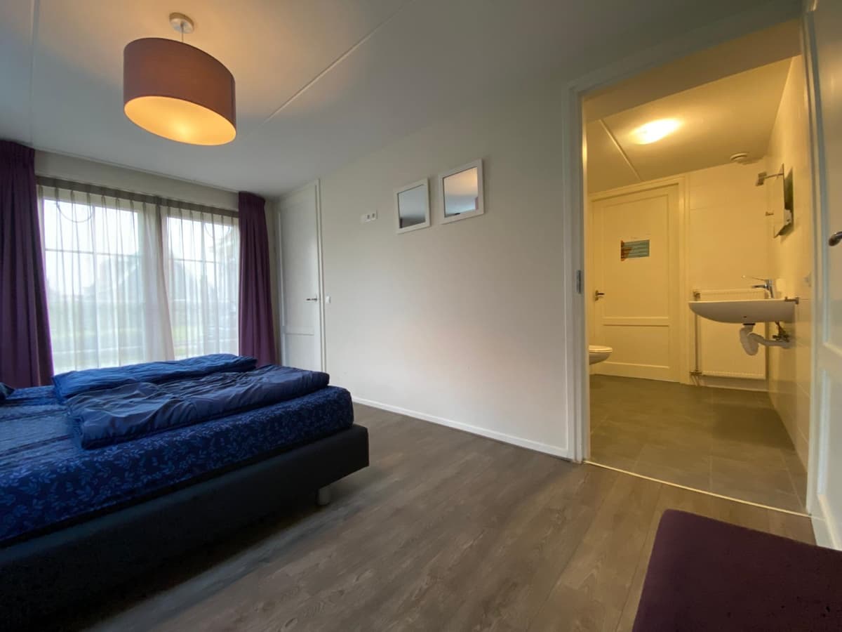Slaapkamer met tweepersoonsbed, raam met gordijnen en aansluiting naar de badkamer met toilet en wastafel.