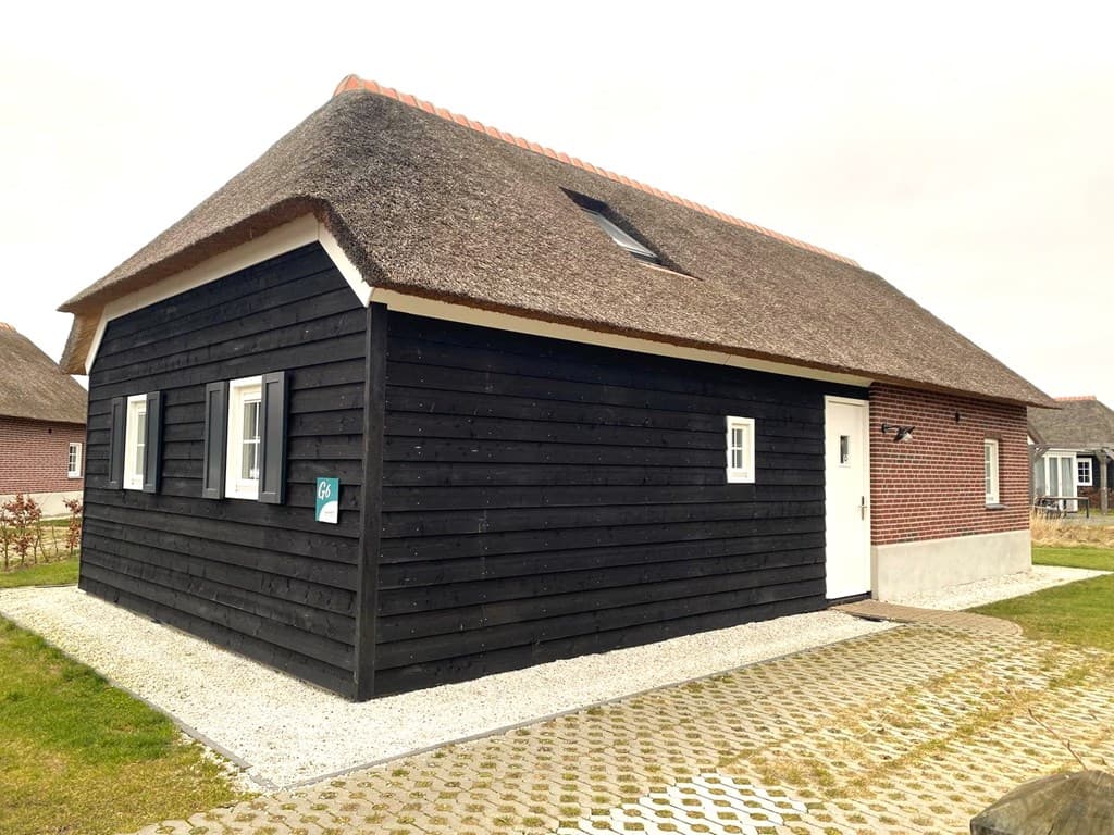 Zwart houten vakantiehuis met rieten dak en een lichte gevel, omringd door gras en grindpaden