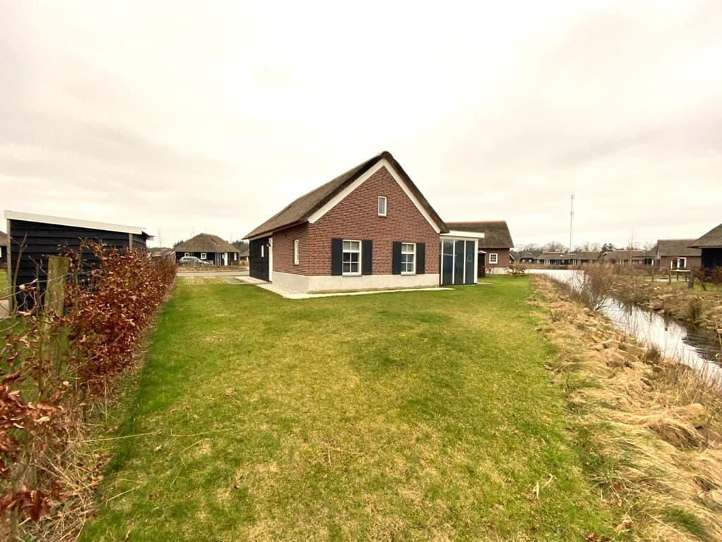 Vakantiewoning met roodbricks en een rieten dak, omgeven door groen gras en waterpartijen.