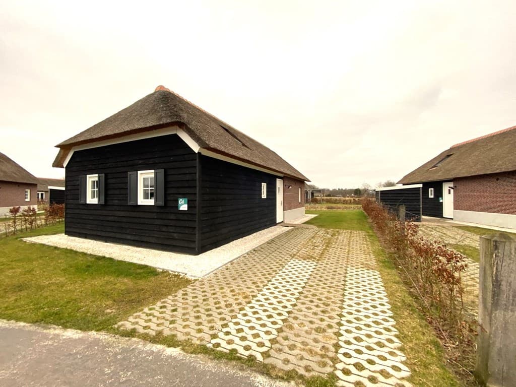 Zwart houten vakantiehuis met rieten dak aan een grindpad, omringd door gras en lage heggen.