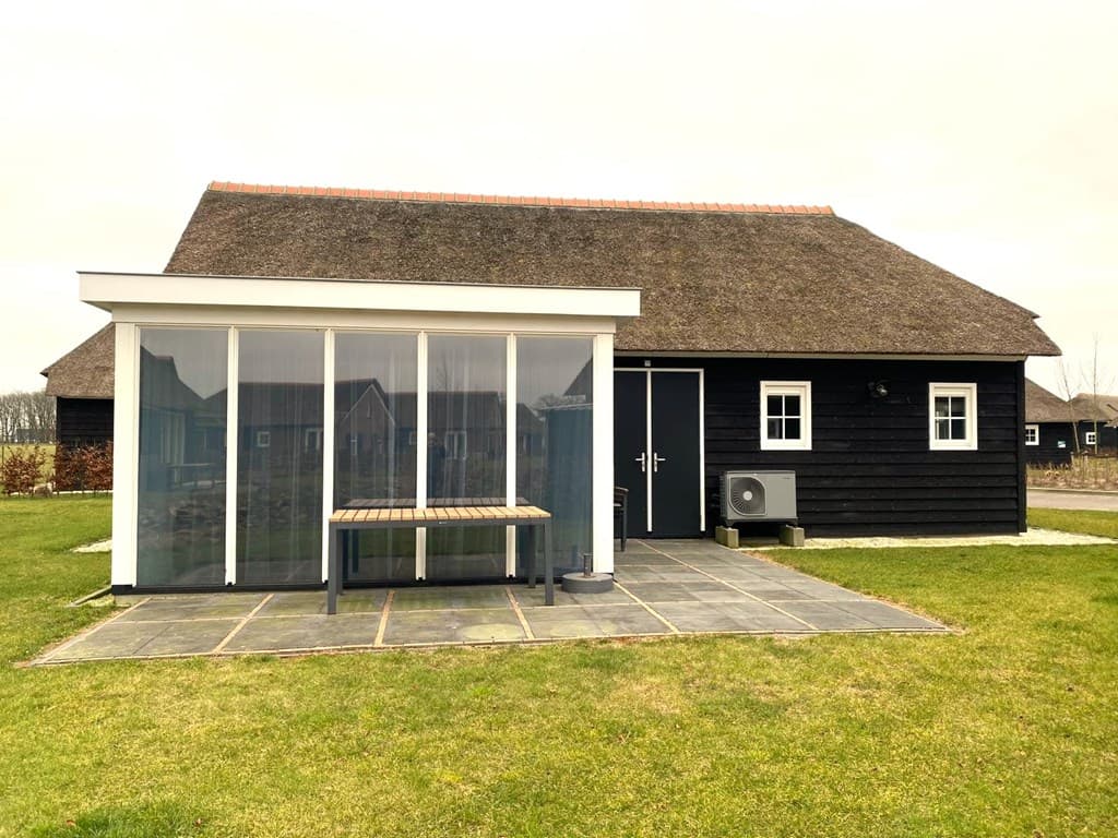 Zwarte vakantiewoning met een rieten dak, terras met glazen wand en een houten tafel voor de deur, gelegen in een groene omge