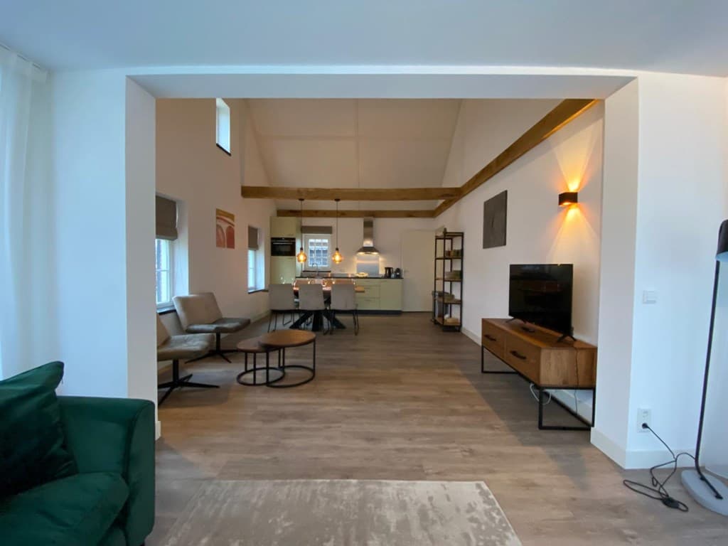 Ruime woonkamer met keuken en dubbele hoogte, voorzien van houten balken en moderne inrichting, twee zithoeken en tv-meubel