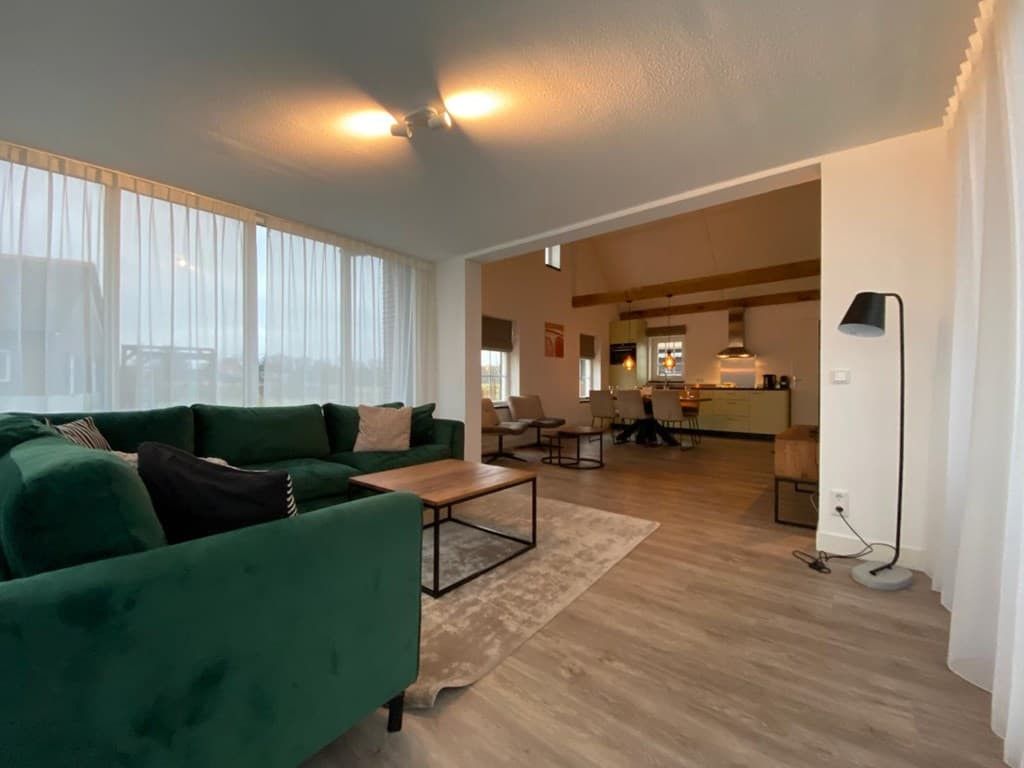 Ruimtelijke woonkamer met groene bank, houten tafel, grote ramen en zicht op open keuken en eetruimte.