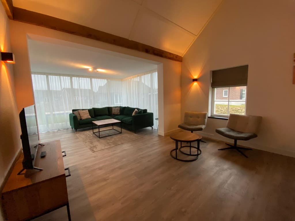 Woonkamer met een groene bank, twee fauteuils en een tv-meubel, grote ramen met lichtdoorlatende gordijnen.