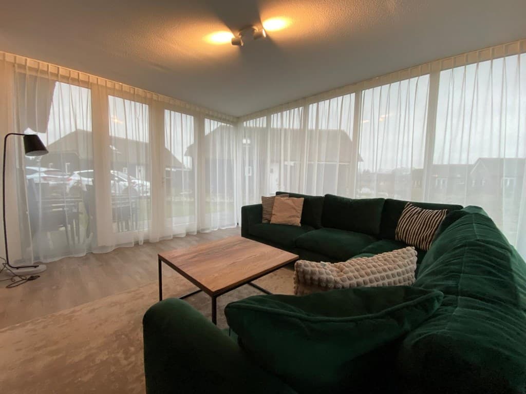 Ruime woonkamer met grote ramen, groene bank, houten salontafel en een vloerlamp in een vakantiehuis.